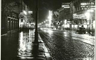 Nachtaufnahme von Bukarest in den 1930-iger Jahren. Eine nasse Straße, in der sich die Lichter der Reklameschilder spiegeln. Autos fahren über die Straße, Menschen stehen vor den hell beleuchteten Lokalen und reden. 