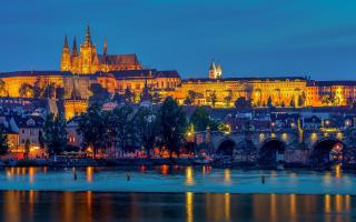 Abendliche Ansicht der Prager Burg und der Karlsbrücke in Prag, Tschechien. Die historischen Gebäude sind golden beleuchtet und spiegeln sich im ruhigen Wasser der Moldau. Die Brücke mit ihren Steinbögen und Statuen verbindet die beiden Ufer der Stadt. Der Himmel zeigt ein tiefes Blau der Dämmerung.